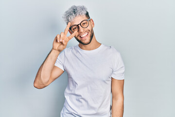 Young hispanic man with modern dyed hair wearing white t shirt and glasses doing peace symbol with fingers over face, smiling cheerful showing victory