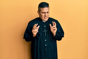 Young latin priest man standing over yellow background disgusted expression, displeased and fearful doing disgust face because aversion reaction.