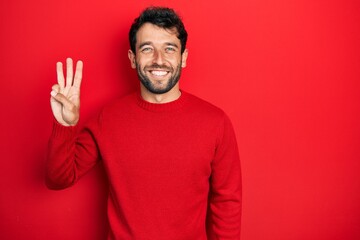 Wall Mural - Handsome man with beard wearing casual red sweater showing and pointing up with fingers number three while smiling confident and happy.