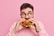 Gluttony and overeating concept. Handsome bearded man bites delicious hamburger feels very hungry looks away wears round spectacles poses against pink background enjoys cheat meal. Unhealthy eating
