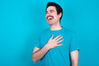 Joyful young handsome Caucasian man with moustache wearing blue t-shirt against blue background expresses positive emotions recalls something funny keeps hand on chest and giggles happily.