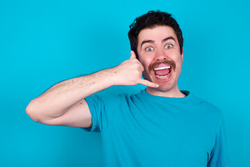 Wall Mural - young handsome Caucasian man with moustache wearing blue t-shirt against blue background makes phone gesture, says call me back again, has glad expression.