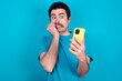 Portrait of pretty frightened young handsome Caucasian man with moustache wearing blue t-shirt against blue background chatting biting nails after reading some scary news on her smartphone.