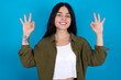 Glad young beautiful tattooed girl standing against blue background shows ok sign with both hands as expresses approval, has cheerful expression, being optimistic.