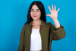 © Jihan - young beautiful tattooed girl standing against blue wall showing and pointing up with fingers number five while smiling confident and happy.