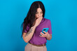 young beautiful tattooed girl wearing blue t-shirt standing against blue background being deeply surprised, stares at smartphone display, reads shocking news on website, Omg, its horrible!