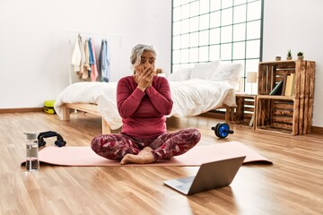 Canvas Print - Middle age woman with grey hair training at home looking at exercise video on laptop shocked covering mouth with hands for mistake. secret concept.