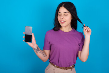 Photo of nice pretty young beautiful tattooed girl wearing purple t-shirt standing against blue background demonstrate phone screen hold hair tails