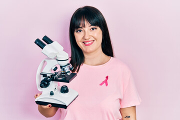 Sticker - Young hispanic woman wearing pink cancer ribbon on shirt holding microscope looking positive and happy standing and smiling with a confident smile showing teeth