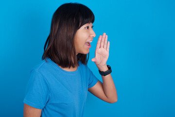 young beautiful asian girl wearing blue t-shirt against blue background look empty space holding hand near her face and screaming or calling someone.