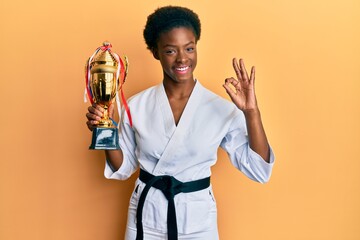 Poster - Young african american girl wearing karate kimono holding trophy doing ok sign with fingers, smiling friendly gesturing excellent symbol