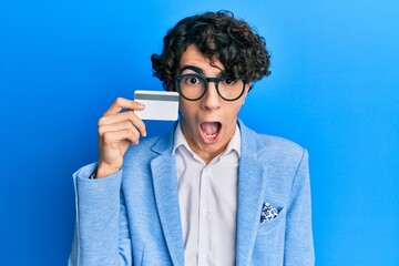 Wall Mural - Hispanic young man holding credit card scared and amazed with open mouth for surprise, disbelief face