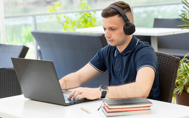 Young man watches a webinar and listens to headphones while sitting at a table in a co working cafe. A male freelancer works at a laptop and listens to music.