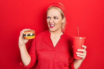 Canvas Print - Young blonde woman eating a tasty classic burger with fries and soda angry and mad screaming frustrated and furious, shouting with anger. rage and aggressive concept.