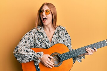 Wall Mural - Young irish woman wearing boho style playing classical guitar angry and mad screaming frustrated and furious, shouting with anger. rage and aggressive concept.