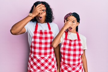 Poster - Beautiful african american mother and daughter wearing baker uniform peeking in shock covering face and eyes with hand, looking through fingers afraid