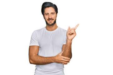 Young hispanic man wearing casual white t shirt with a big smile on face, pointing with hand and finger to the side looking at the camera.