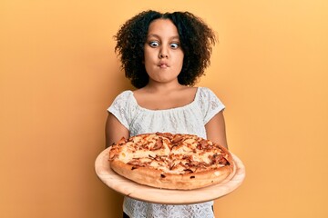 Sticker - Young little girl with afro hair holding italian pizza making fish face with mouth and squinting eyes, crazy and comical.