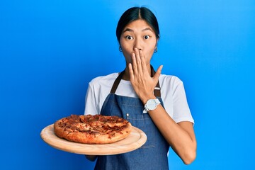 Wall Mural - Young chinese woman wearing waiter apron holding italian pizza covering mouth with hand, shocked and afraid for mistake. surprised expression