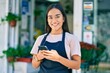 Young latin shopkeeper girl smiling happy using smartphone at florist.