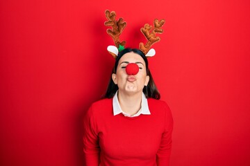 Poster - Young hispanic woman wearing deer christmas hat and red nose looking at the camera blowing a kiss on air being lovely and sexy. love expression.