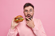 Shocked bearded adult man holds delicious hamburger eats fast food has unhealthy nutrition holds chin dressed in casual jumper isolated over pink background. Hungry guy with appetizing burger