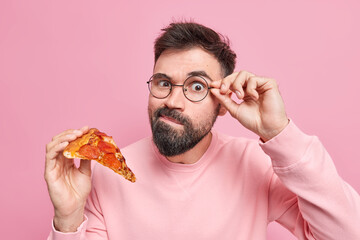 Harmful delicious food. Handsome bearded man has tasty snack keeps hand on rim of spectacles holds slice of appetizing italian pizza has harmful unhealthy eating habits isolated on pink wall