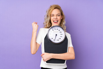 Poster - Young blonde woman isolated on purple background with weighing machine and doing victory gesture