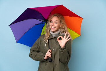 Poster - Young blonde woman holding an umbrella isolated on blue background showing ok sign with fingers