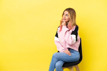 Wall Mural - Young woman sitting on a chair over isolated yellow background having doubts