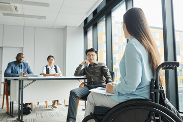 Wall Mural - Side view portrait of young woman in wheelchair giving speech to diverse business team in office, copy space