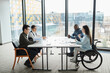 © Seventyfour - Wide angle view at diverse business team discussing project at table in modern office with focus on young woman in wheelchair sharing ideas in foreground, copy space