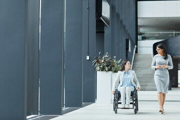 Wall Mural - Full length portrait of young businesswoman in wheelchair talking to female colleague while moving towards camera in office lobby, copy space