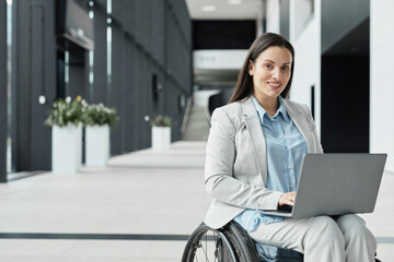 Portrait of successful young businesswoman in wheelchair using laptop and smiling at camera in office lobby, copy space