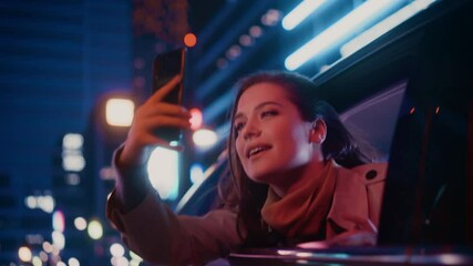 Wall Mural - Excited Female is Looking Out of the Window from Backseat of a Car at Night. Woman Taking Photos and Videos on a Smartphone with Amazement of How Beautiful is the City Street with Working Neon Signs.