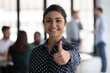 © fizkes - Photo portrait of happy Indian female business leader gesturing like, showing thumb up. Team working in background. Satisfied client woman gives approval positive feedback, recommends company service