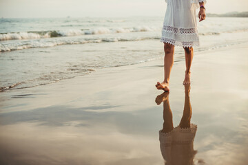 a middle-aged woman walks relaxed along the seashore. she wears a white thread dress. the light is golden from sunset.