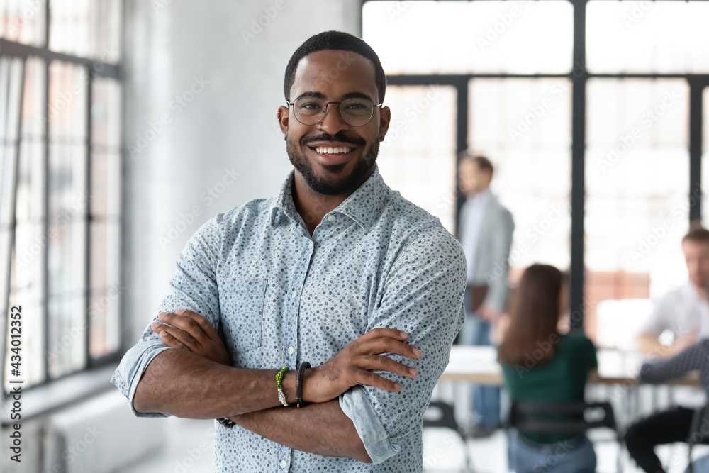 Portrait of happy African American small business owner posing with hands folded. Millennial black male team leader smiling, looking at camera, employees working in modern office behind. Head shot - obrazy, fototapety, plakaty 
