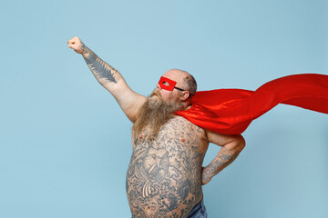 Side view fun emotive fat pudge obese chubby overweight blue-eyed bearded man 30s in red cartoon character cloak mask naked torso keeps arm in flying gesture isolated on pastel blue background studio.