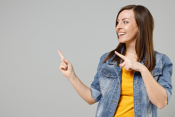 Wall Mural - Young smiling happy caucasian woman 20s wearing casual trendy denim jacket yellow t-shirt pointing index finger aside on workspace copy space area mock up isolated on grey background studio portrait