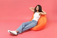 Young Fun Smiling Happy Friendly Positive Cute African American Woman 20s Wear Casual White Tank Shirt Sitting In Orange Bean Bag Chair Hold Hands Behind Neck Head Isolated On Pink Color Background
