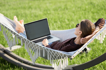 Wall Mural - Young woman using laptop outdoor. Young beautiful girl lying in a hammock and working on computer. Freelance, summer, enjoy life, student lifestyle, distance studying and online learning concept