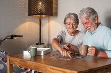 Smiling Senior Couple Doing A Jigsaw Puzzle At Home On Wooden Table. Vintage Bicycle In The Corner
