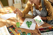 © New Africa - Young woman drawing leaf with watercolors at table indoors, closeup