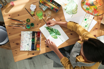 Canvas Print - Young woman drawing leaf with watercolors at table indoors, top view