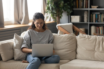 Wall Mural - Happy young woman relax on couch at home browse wireless internet on laptop. Smiling millennial female in glasses work online on computer, talk speak on video call. Technology concept.