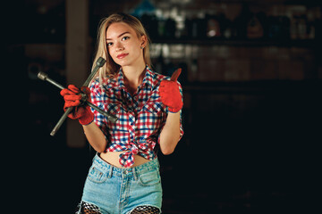 A girl in a bright plaid shirt and short denim shorts holds a balloon wrench at the service station. Handsome mechanic at work. Portrait of a beautiful model in a car garage.