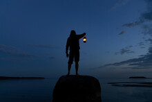 Man Standing On The Rock And Holding Old Lantern Outdoors Near The Sea At Night.  Light And Hope Concept.
