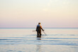 A man with a metal detector and scoop searches for treasures in the sea.