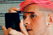 © Luis Mario Hernandez/ADDICTIVE STOCK - Homosexual man with piercings and modern haircut applying mascara on eyelashes with applicator against cellphone